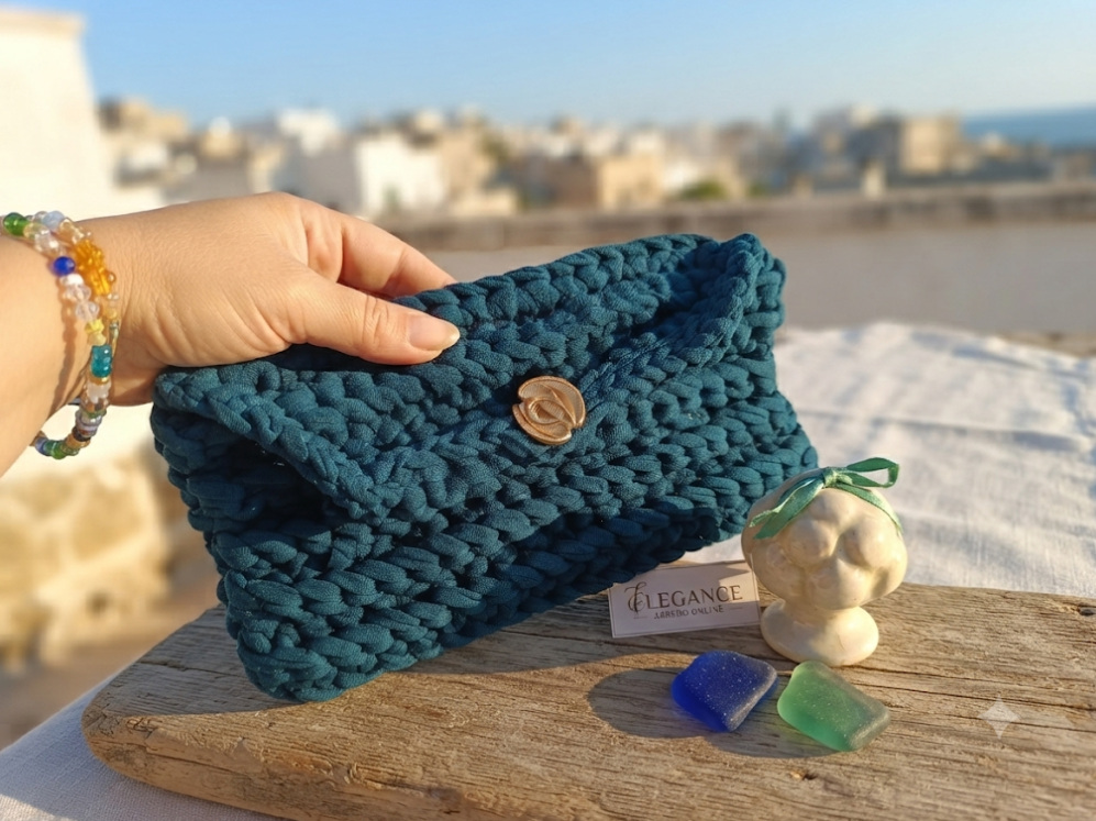 Pochette Ottanio vista frontale con bottone dorato e logo Elegance Arredo, su tavolo in legno con vista sui tetti bianchi