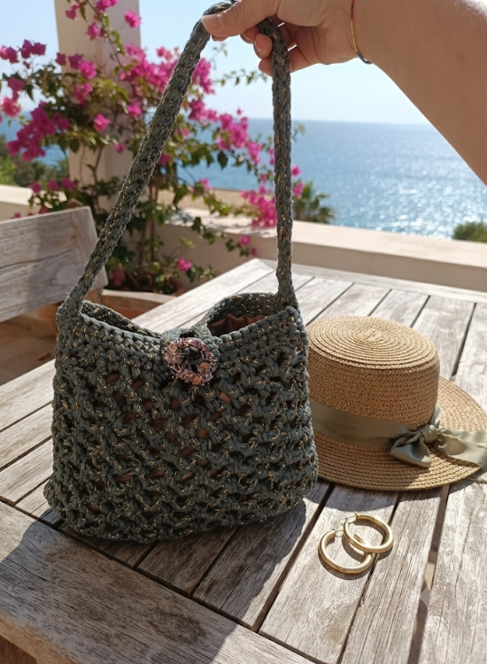 Borsa Verde Traforata all'uncinetto in tulle iridescente e filo dorato con chiusura gioiello a coroncina floreale, su tavolo in legno con vista mare