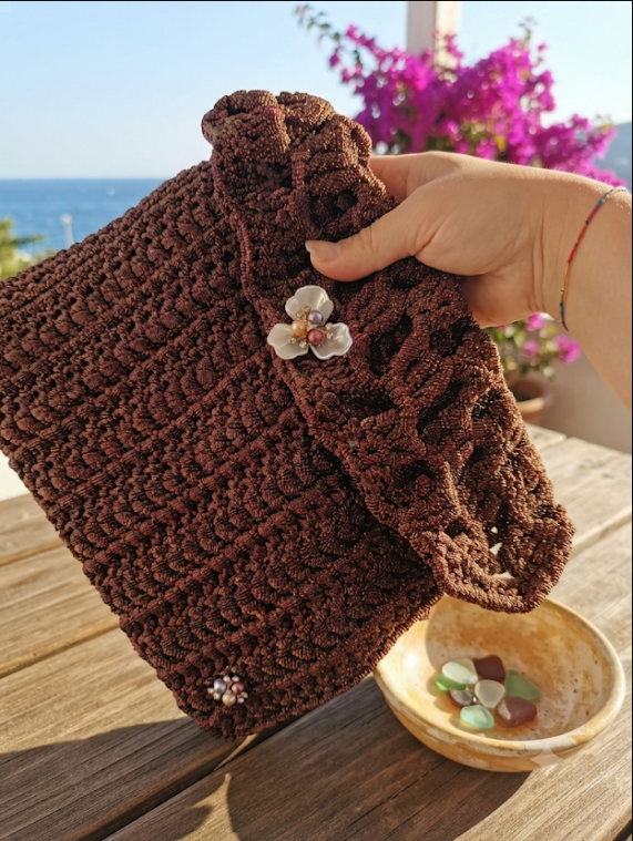 Pochette Bronzo all'uncinetto in tulle iridescente con chiusura a fiore d'orchidea madreperlato e dettagli in perle, su tavolo in legno con vista mare